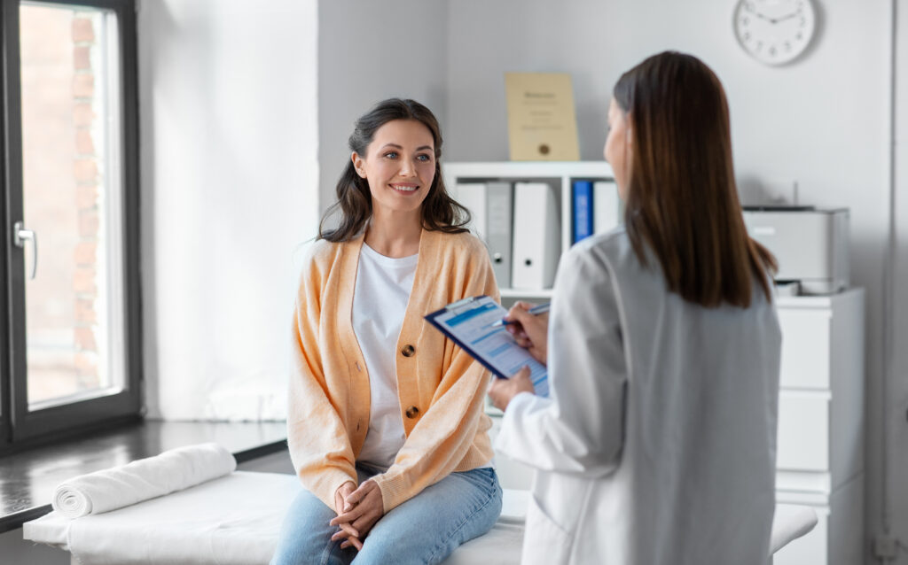 Patient seated on exam table speaking with clinician holding a clipboard