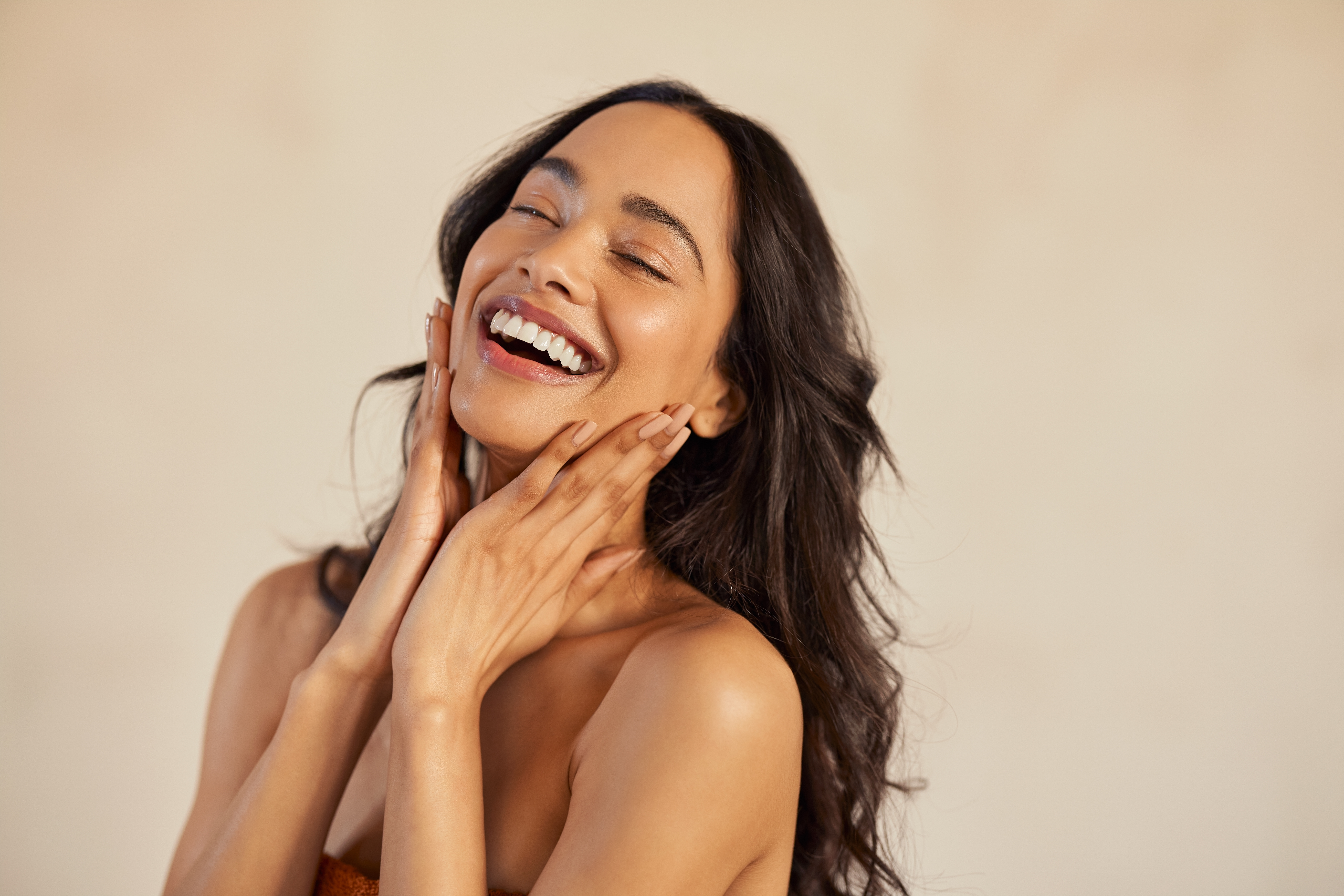 Woman with eyes closed touching her jaw and cheeks against a neutral background
