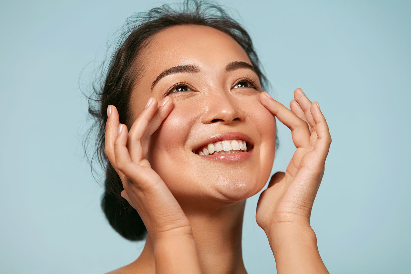 Woman smiling with hands placed on her cheeks against a light blue background.