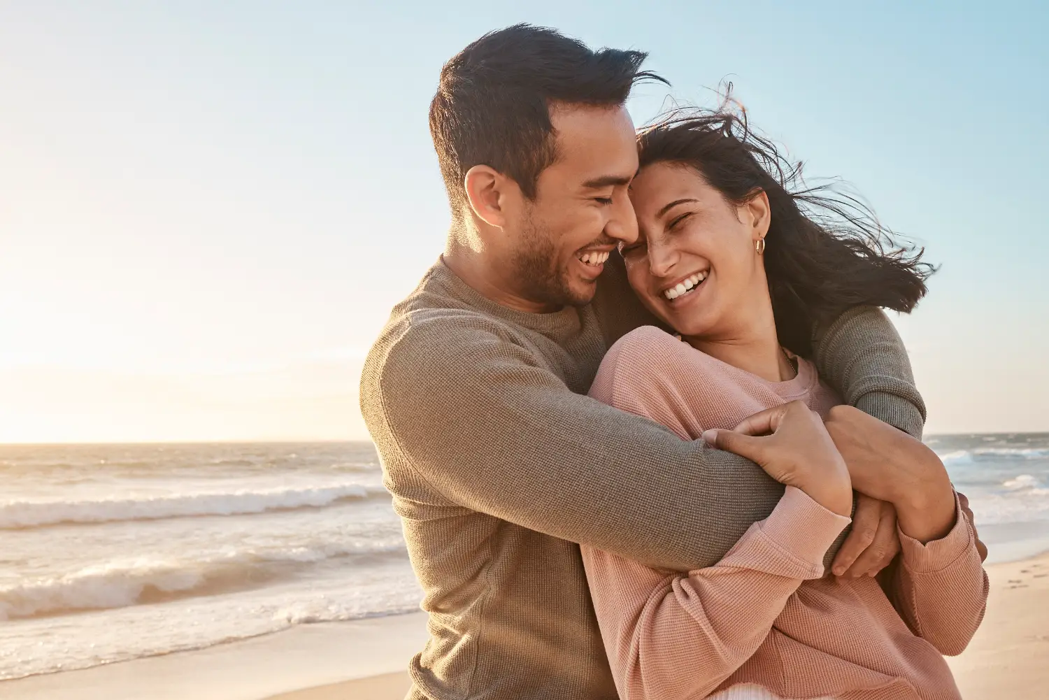 Smiling couple hugging each other on a beach with ocean waves in the background at sunset.
