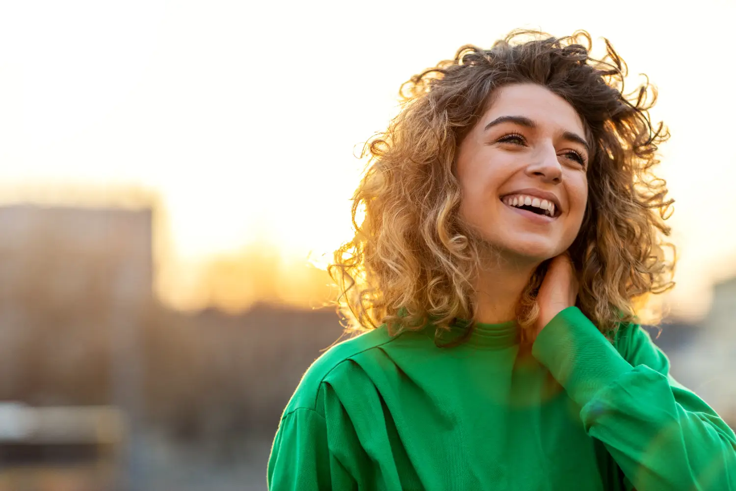 Woman with curly hair smiling outdoors at sunset while touching her neck.