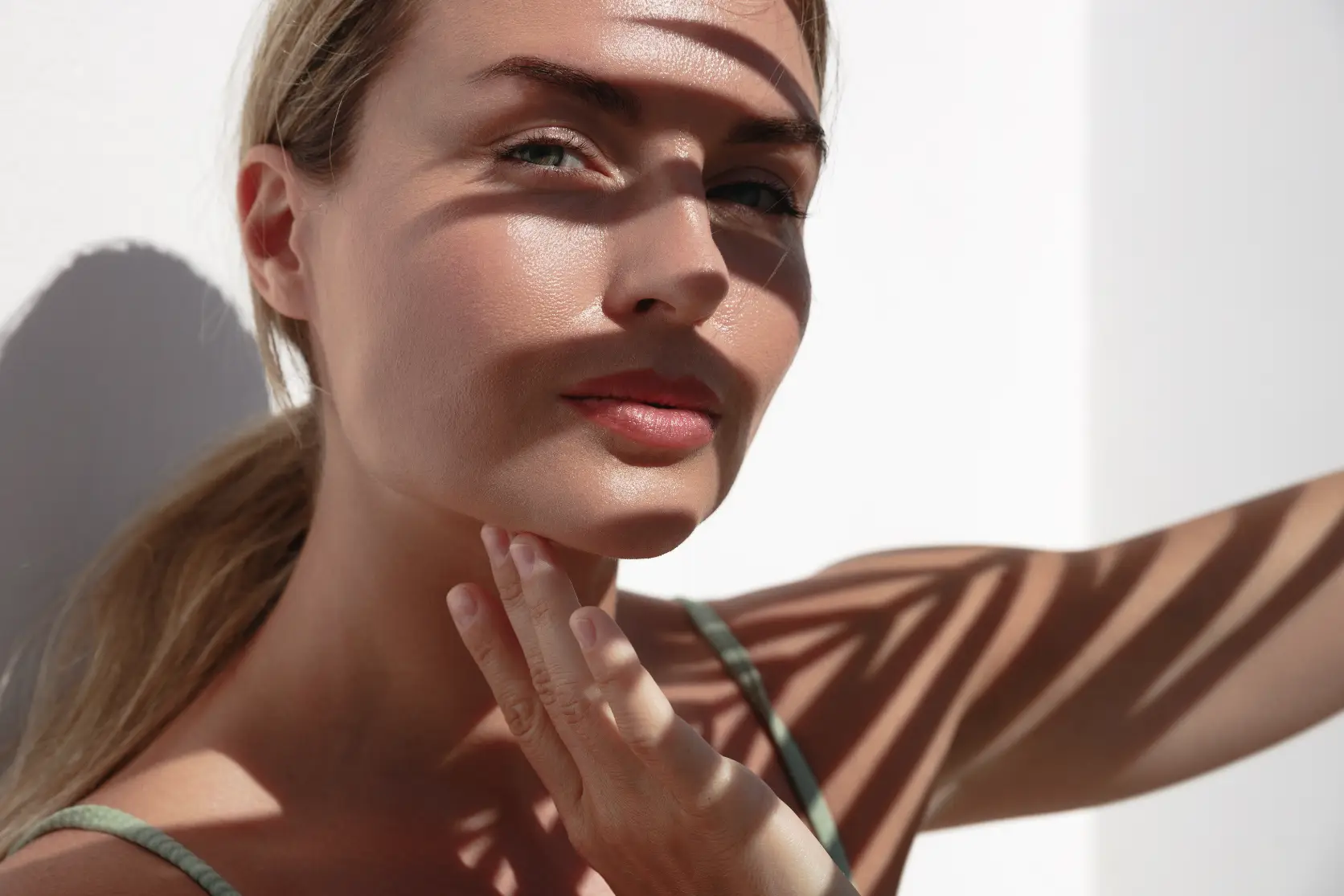 Woman with glowing skin touching her jawline in sunlight with shadow patterns across her face